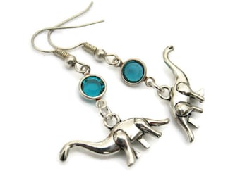 May include: A pair of silver dangle earrings featuring a turquoise blue crystal and a dinosaur charm.