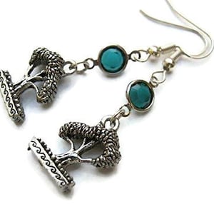May include: Silver-toned dangle earrings featuring a tree design and teal-colored gemstones. The earrings have a hook closure and are set against a white background. The tree design is detailed and textured.