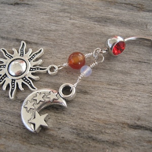 May include: Silver belly button ring with a red gem, a sun charm, and a crescent moon charm with stars. The charms are attached to the ring with silver wire and beads.