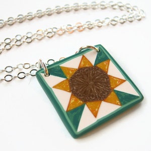May include: A silver chain necklace with a square pendant featuring a brown and yellow sunflower design on a white background with a teal border.