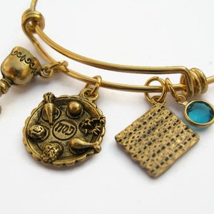 May include: Gold charm bracelet with four charms: a Seder plate, a matzah, a goblet, and a blue crystal. The charms are attached to a gold bangle bracelet.