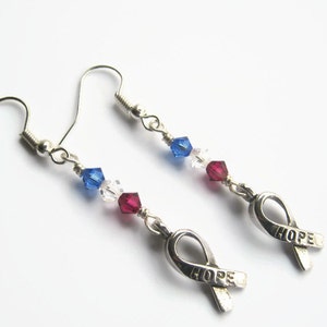 May include: Silver earrings with a red, white, and blue crystal bead design. The earrings feature a silver ribbon charm with the word "HOPE" engraved on it.