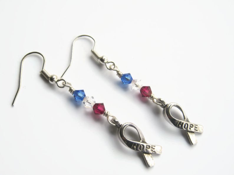 May include: Silver earrings with a red, white, and blue crystal bead design. The earrings feature a silver ribbon charm with the word "HOPE" engraved on it.