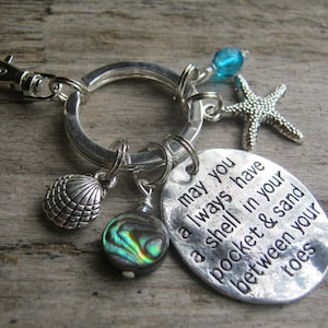 May include: Silver keychain with a charm that reads "may you always have a shell in your pocket & sand between your toes". The keychain also features a seashell, a starfish, a blue bead, and a mother-of-pearl bead.