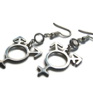 May include: A pair of silver earrings with a clear crystal and a transgender symbol charm.