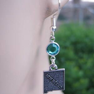 May include: Silver earrings with a turquoise blue crystal and a silver charm shaped like the state of New Mexico with the words "New Mexico" inscribed on it.