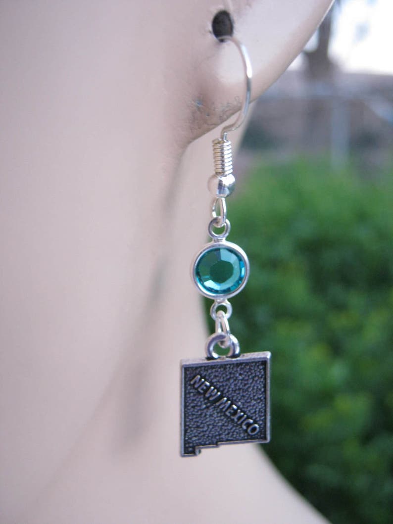 May include: Silver earrings with a turquoise blue crystal and a silver charm shaped like the state of New Mexico with the words "New Mexico" inscribed on it.