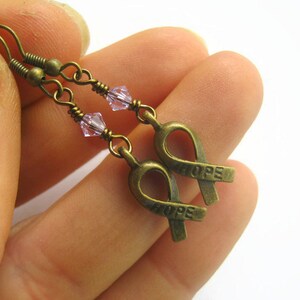 Puede incluir: Un par de pendientes de bronce con un dije de cristal rosa y un dije de cinta de bronce con la palabra "HOPE" grabada.