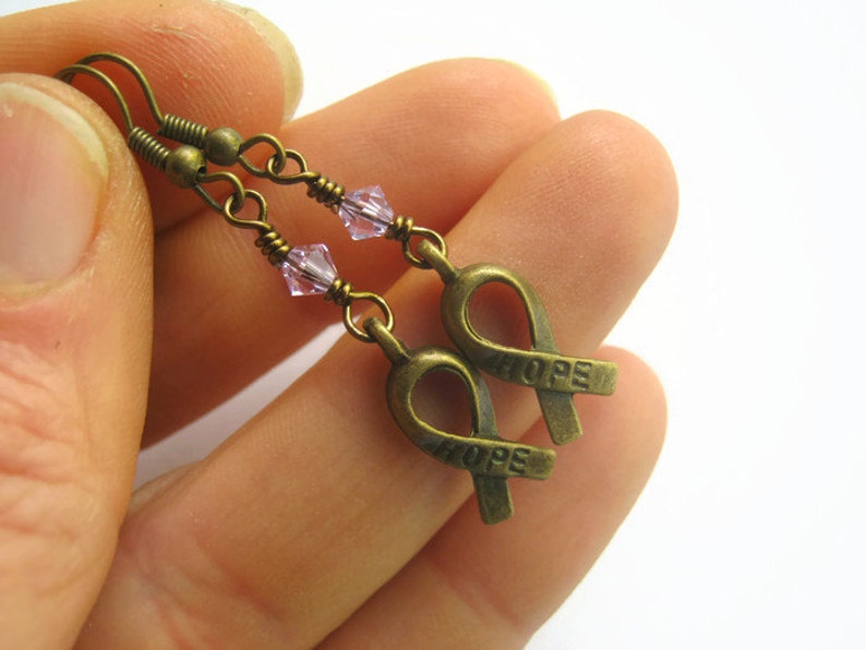 Puede incluir: Un par de pendientes de bronce con un dije de cristal rosa y un dije de cinta de bronce con la palabra "HOPE" grabada.
