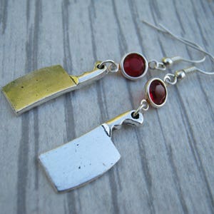 May include: A pair of silver earrings featuring a miniature cleaver charm and a red rhinestone.