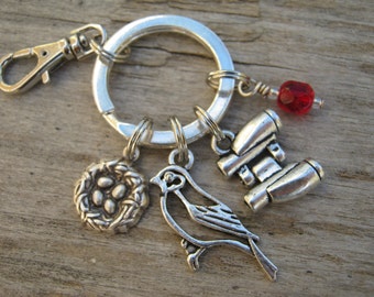 May include: Silver keychain with a bird, a nest with eggs, binoculars, and a red bead charm.