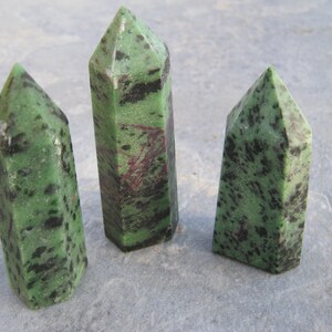 Puede incluir: Tres puntas de piedra preciosa verde, negra y rosa, también conocidas como zoisita, están dispuestas en fila sobre una superficie gris.