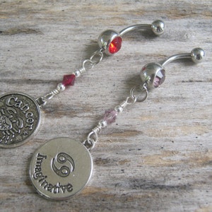 May include: Two silver belly button rings with charms. The charms are round and feature the zodiac symbol for Cancer. One charm says "Cancer" and the other says "Imaginative".