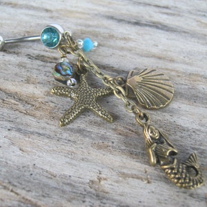 May include: A silver body piercing with a blue gem and a charm chain featuring a mermaid, a starfish, a seashell, and iridescent beads.