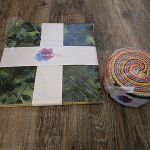 May include: A stack of fabric squares with a green floral pattern, wrapped with a white band and the "boundless fabrics" logo. Next to it is a roll of colorful fabric strips, also labeled with the "boundless fabrics" logo.