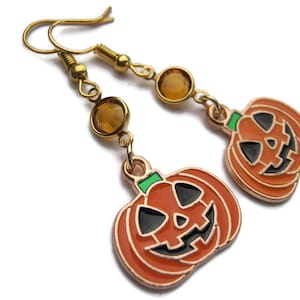 May include: Gold-tone Halloween earrings featuring orange jack-o'-lantern charms with black facial features and green stems. Each earring has a dangling amber-colored gem. The earrings are designed for pierced ears.
