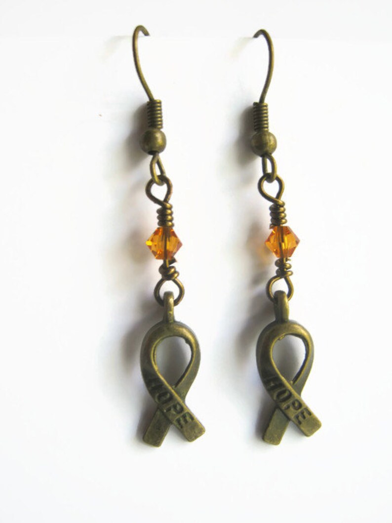 Puede incluir: Un par de pendientes de bronce con un peque&ntilde;o cristal &aacute;mbar y un dije de cinta de bronce con la palabra "HOPE" grabada.