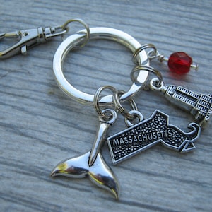 May include: Silver keychain with a red bead, a whale tail charm, a charm of the state of Massachusetts, and a charm of a building.