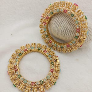 May include: Two circular gold-toned bangles featuring an intricate metalwork design. Each bangle is adorned with small white faux pearls and tiny floral accents in shades of red, green, and light blue, arranged in a repeating pattern around the band.