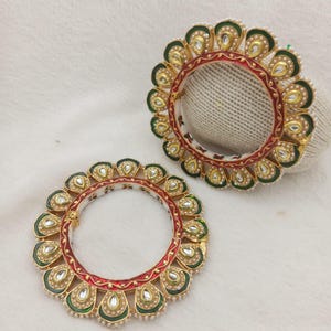 May include: Two circular bangles featuring a central red band bordered by gold metal. The outer edge is decorated with a series of teardrop-shaped motifs in green and gold, each containing a white stone, accented by small white beads along the perimeter.