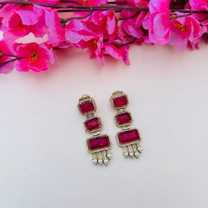 May include: A pair of gold-tone drop earrings featuring three rectangular, deep pink stones arranged vertically. Each stone is framed by small clear crystals, and the bottom of each earring is finished with a row of dangling, teardrop-shaped clear crystals.