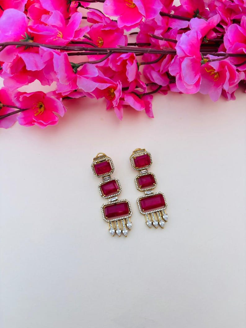 May include: A pair of gold-tone drop earrings featuring three rectangular, deep pink stones arranged vertically. Each stone is framed by small clear crystals, and the bottom of each earring is finished with a row of dangling, teardrop-shaped clear crystals.