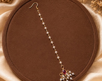 May include: A gold-toned maang tikka headpiece featuring a long chain adorned with small white pearls. The piece terminates in a teardrop-shaped pendant with a deep red stone at the centre, surrounded by a cluster of dangling white pearl beads.