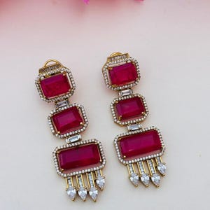 May include: A pair of gold-tone drop earrings featuring three stacked rectangular deep pink stones. Each stone is framed by small clear crystals. The bottom of each earring has a fringe of small clear baguette and pear-shaped crystals.