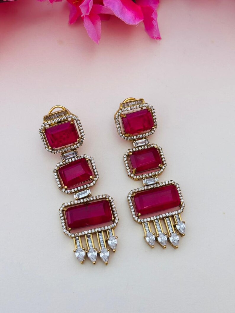 May include: A pair of gold-tone drop earrings featuring three stacked rectangular deep pink stones. Each stone is framed by small clear crystals. The bottom of each earring has a fringe of small clear baguette and pear-shaped crystals.