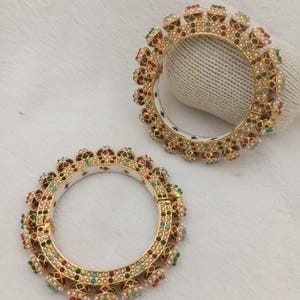 Peut inclure: Deux bracelets joncs dorés avec un intérieur blanc et un extérieur orné de petites pierres multicolores rouges, vertes et bleues, agrémentées de minuscules perles blanches disposées en un motif répétitif sur toute la circonférence.