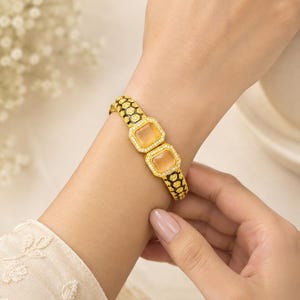 Pode incluir: Uma pulseira de tom dourado com duas pedras quadradas de cor pêssego no centro. A banda apresenta um padrão de favo de mel preto com contornos dourados e é decorada com pequenas pedras transparentes e brilhantes por todo o design.