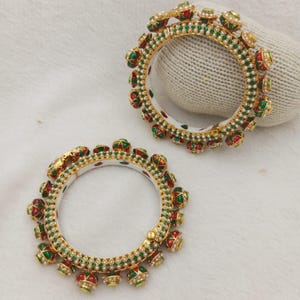 Può includere: Due bracciali circolari con base in metallo color oro e dettagli intricati in smalto rosso e verde. I bracciali sono decorati con piccole perle bianche lungo i bordi interni ed esterni e presentano elementi decorativi pendenti.
