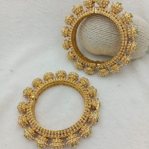 Può includere: Un paio di bracciali rigidi color oro caratterizzati da un intricato design in filigrana metallica. Ogni bracciale circolare è decorato con una serie di piccoli ciondoli sferici lungo il bordo esterno, arricchiti da minuscole perline.
