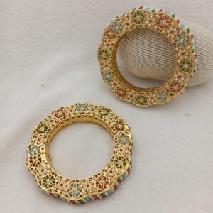 May include: Two gold-toned bangles featuring a scalloped edge design. Each bangle is decorated with a repeating floral pattern inlaid with small red, green, and blue stones, accented by tiny white beads set within the gold metal framework.
