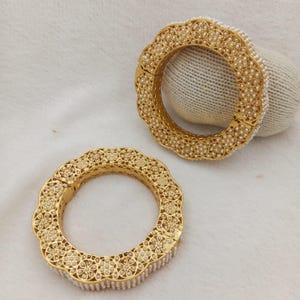 May include: Two gold-toned bangles featuring a scalloped edge design. Each bangle is intricately decorated with a repeating floral pattern filled with small white beads. The metal surface has a polished finish, and the circular structure is consistent.