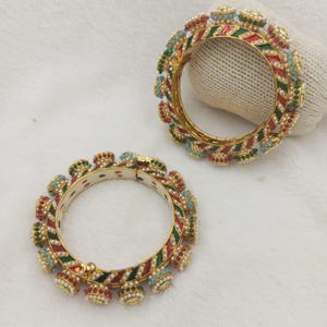 Può includere: Due bracciali rigidi dorati con strisce diagonali in smalto rosso e verde. I bordi esterni sono decorati con piccoli ciondoli a forma di campanella, arricchiti da minuscole perle bianche e pietre di colore rosso, verde e azzurro.
