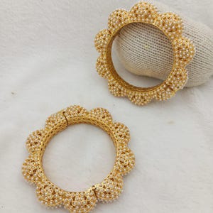 Pode incluir: Dois braceletes dourados com um design de borda festonada em forma de flor. Cada bracelete é detalhado com um padrão de grade metálica texturizada e incrustado com inúmeras pequenas pérolas brancas cobrindo toda a superfície externa.