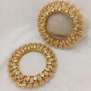 May include: Two circular gold-toned bangles featuring a repeating pattern of teardrop-shaped white stones and small white beads. The metal surface is textured with intricate detailing, and each bangle has a hinged opening mechanism.