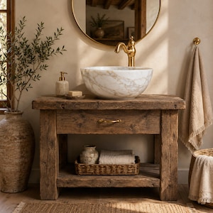 Puede incluir: Un mueble de baño de madera rústica con un lavabo de mármol blanco y grifo dorado, un cajón con tirador dorado y un estante inferior abierto con una cesta tejida, un tarro de cerámica y un paño doblado, además de una toalla beige colgada.