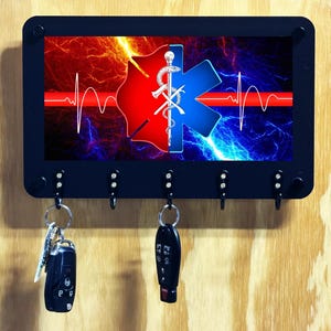 May include: A rectangular black wall-mounted key holder with five metal hooks. The center graphic features a split red and blue design with a central Star of Life symbol, a white EKG heartbeat line, and a lightning-like background effect.