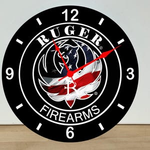 May include: A round wall clock with a black face, white numerals at 12, 3, 6, and 9, and white tick marks. The center features the Ruger Firearms logo overlaid on a waving American flag. The clock has two thin, bright red hands for timekeeping.