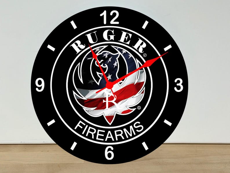 May include: A round wall clock with a black face, white numerals at 12, 3, 6, and 9, and white tick marks. The center features the Ruger Firearms logo overlaid on a waving American flag. The clock has two thin, bright red hands for timekeeping.