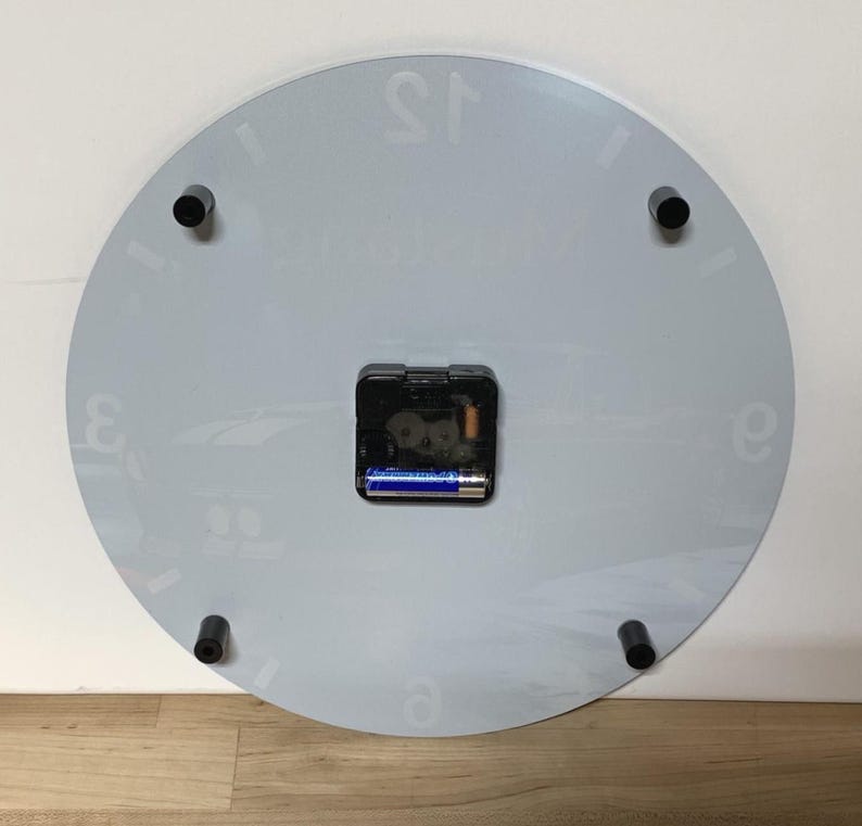 May include: The reverse side of a circular wall clock featuring a light gray surface with faint white numerals. A black battery-operated quartz movement mechanism is centered on the back, holding a single blue and silver AA battery in place.