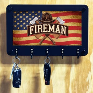 May include: A rectangular black wall-mounted key holder featuring a graphic of the American flag, a firefighter helmet, and two crossed axes. The word FIREMAN is printed in large white letters across the center. Five metal hooks hold two key fobs.