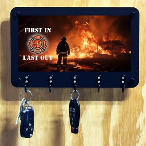 May include: A black rectangular wall-mounted key holder featuring a graphic of a firefighter standing before a large fire. The text FIRST IN and LAST OUT surrounds a central fire rescue emblem. Five black hooks hold two sets of car keys.