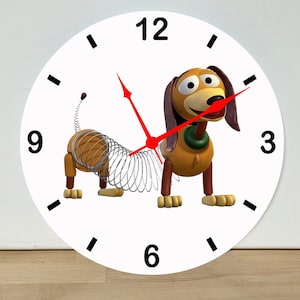 May include: A round white wall clock featuring a graphic of a brown dachshund toy with a metal spring body. The clock face displays black numbers at the 3, 6, 9, and 12 positions, with black tick marks for other hours and bright red clock hands.