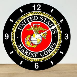 May include: A round wall clock featuring the United States Marine Corps emblem in the center. The clock face has a black background with white numbers 3, 6, 9, and 12, and white hour, minute, and second hands. The emblem includes a red circle.