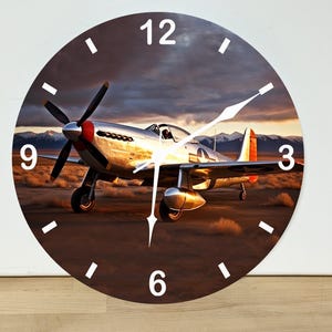 May include: A round wall clock featuring a photographic print of a silver P-51 Mustang fighter plane parked on a desert runway at sunset. The clock face has white hour markers and white hands, with the numbers 3, 6, 9, and 12 printed in white.