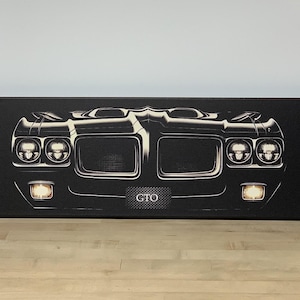 May include: A rectangular canvas wall art print featuring a high-contrast, black and white front-facing illustration of a classic muscle car. The design highlights the vehicle's dual headlights, grille, and bumper, with the text GTO centered below.