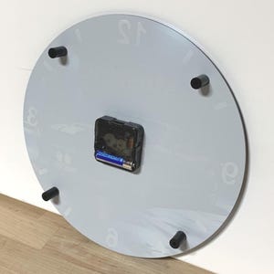 May include: A circular wall clock viewed from the back, featuring a light gray surface with faint numeral markings. A black plastic clock mechanism is centered on the back, holding a single AA battery. Four black cylindrical spacers are attached.